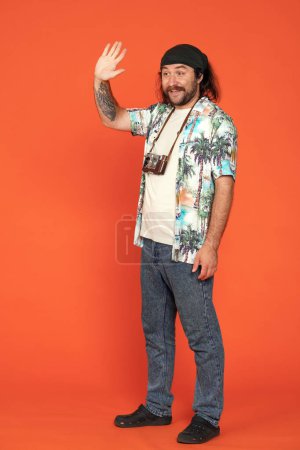 Téléchargez les photos : Voyageur masculin en studio sur fond orange. Un homme avec une caméra autour du cou leva la main pour saluer. Plan vertical - en image libre de droit