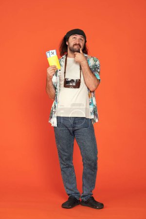 Photo for Male traveler holding his passport with airline tickets and thinking about something with his hand to his chin. Man with full length retro camera in studio on orange background. Vertical shot. Air - Royalty Free Image