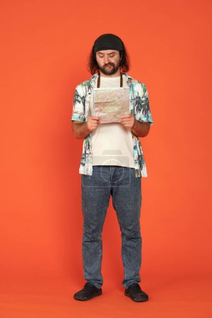 Photo for A male traveler studying a map of an area. A puzzled man is working out a route. Man with camera around his neck in studio on orange background full length. Vertical shot. Hiking and navigation - Royalty Free Image
