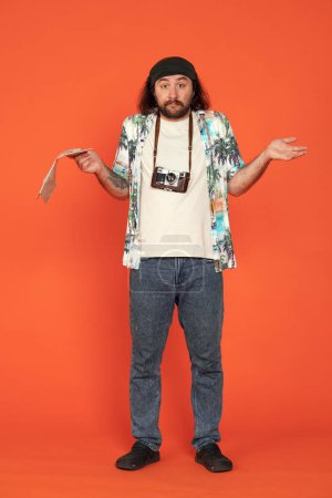 Photo for Confused lost male traveler with map of area, shrugs and spreads arms apart. Man with camera around his neck in studio on orange background in full height. Vertical shot. Hiking and navigation concept - Royalty Free Image