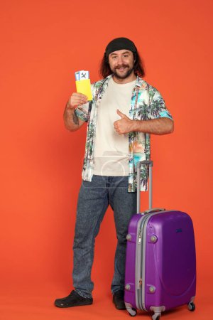 Photo for A male traveler leaning on the handle of a suitcase holding his passport with airline tickets and showing a thumbs up. Happy man in studio on orange background full length. Vertical shot. Air travel - Royalty Free Image