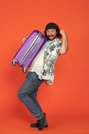 Foto de Un hombre sostiene una maleta grande de color lila y hace un gesto de victoria. Hombre viajero en estudio sobre fondo naranja en altura completa. Disparo vertical - Imagen libre de derechos