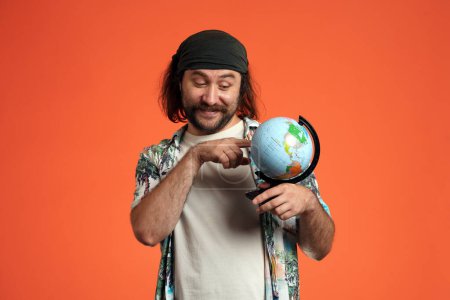 Téléchargez les photos : Un voyageur masculin heureux tient un globe, une carte du monde, en choisissant où aller. L'homme indique un endroit sur la carte. Portrait de l'homme tenant globe en studio sur fond orange de près. Air - en image libre de droit