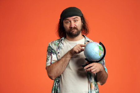 Téléchargez les photos : Un voyageur masculin tient un globe, une carte du monde, en choisissant où aller. Un homme mécontent indique un endroit sur la carte. Portrait d'un homme frustré en studio sur fond orange gros plan - en image libre de droit