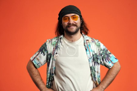 Foto de Retrato de un viajero masculino con gafas de sol en estudio sobre fondo naranja de cerca. Hombre, sonriendo mirando hacia adelante, poniendo sus manos en su cinturón. Vacaciones de verano - Imagen libre de derechos