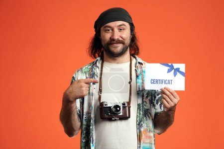 Téléchargez les photos : Heureux voyageur masculin avec appareil photo pointant index à certificat-cadeau dans sa main. Un homme dans un studio sur fond orange gros plan - en image libre de droit