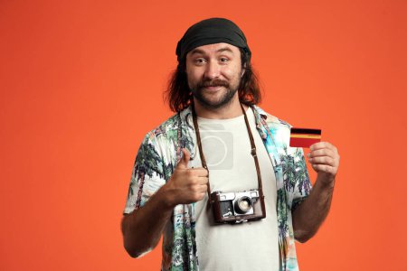 Foto de Hombre viajero sosteniendo una tarjeta de crédito y mostrando los pulgares hacia arriba. Retrato de un hombre con una cámara alrededor del cuello en un estudio sobre un fondo naranja de cerca - Imagen libre de derechos