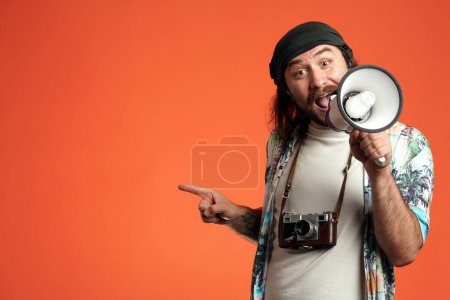 Téléchargez les photos : Un voyageur masculin avec un mégaphone pointe quelque chose avec son index sur le côté. Un homme avec une caméra autour du cou dans un studio sur un fond orange gros plan. Concept de voyage - en image libre de droit