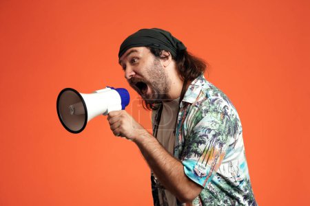 Téléchargez les photos : Un homme tient un mégaphone et y parle fort en criant. Voyageur masculin en studio sur fond orange de près. Concept de voyage - en image libre de droit