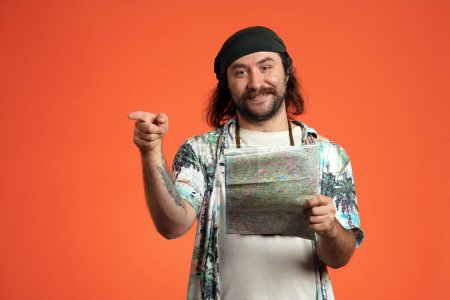 Photo for A man with a map of the area in his hand points his index finger to the side, showing the direction. Male traveler, tourist in the studio on an orange background close up. Hiking and navigation - Royalty Free Image