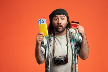 Foto de Retrato de un hombre sorprendido sosteniendo una tarjeta de crédito y pasaporte con billetes de avión. Un hombre con una cámara en el cuello en un estudio sobre un fondo naranja de cerca - Imagen libre de derechos