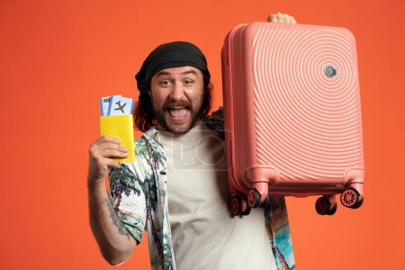 Photo for Happy man holding pink suitcase and passport with air tickets. A man in a studio on an orange background close up. Air flight, journey concept - Royalty Free Image