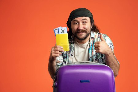 Foto de Un hombre tiene un pasaporte con billetes de avión y muestra un pulgar hacia arriba. Viajero masculino con una maleta lila en el estudio sobre fondo naranja de cerca - Imagen libre de derechos