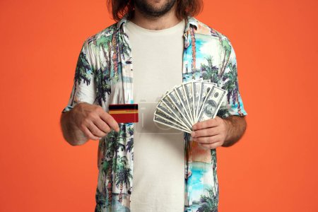 Foto de Un hombre tiene un montón de billetes de dólar y una tarjeta de crédito en su mano de cerca. Hombre viajero en estudio sobre fondo naranja - Imagen libre de derechos