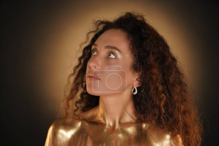 Photo for Beauty and glamour creative advertisement concept. Portrait of female model in studio. Attractive young woman with curly hair and golden makeup looking up, chest coated in golden liquid paint. - Royalty Free Image