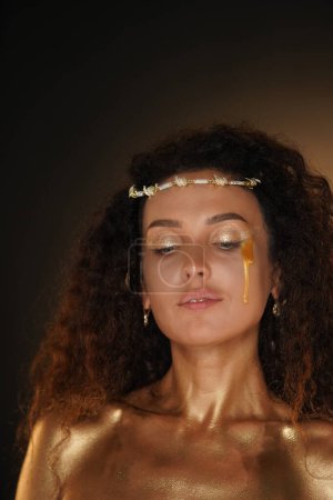 Photo for Beauty and glamour creative advertisement concept. Portrait of female model in studio. Attractive girl wearing tiara and makeup covered in golden paint posing at camera with golden tear on cheek. - Royalty Free Image