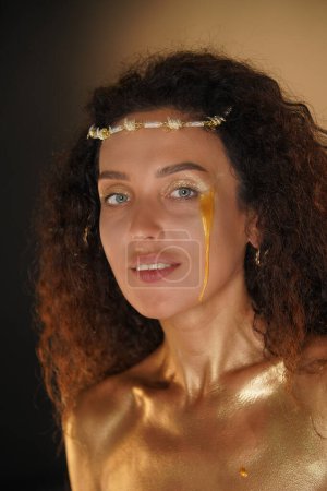 Photo for Beauty and glamour creative advertisement concept. Portrait of female model in studio. Attractive girl wearing tiara and makeup covered in golden paint smiles at camera with golden tear on her skin. - Royalty Free Image
