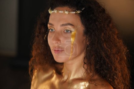 Photo for Beauty and glamour creative advertisement concept. Portrait of female model in studio. Appealing girl wearing tiara and bright makeup covered in golden paint with golden tear flowing down her face. - Royalty Free Image