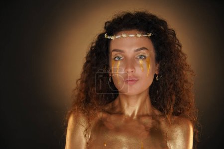 Téléchargez les photos : Concept publicitaire créatif beauté et glamour. Portrait de mannequin femme en studio. Attrayant fille portant la dîme et lumineux maquillage épaules zone couverte de peinture dorée, en regardant la caméra. - en image libre de droit