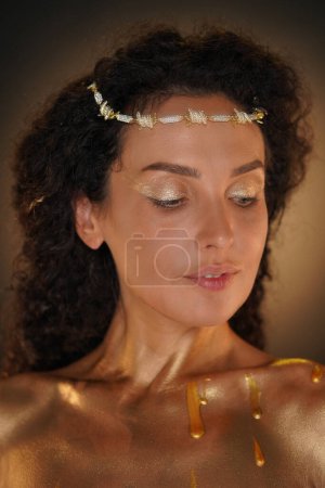 Photo for Beauty and glamour creative advertisement concept. Portrait of female model in studio. Close up shot of attractive girl wearing tiara and elegant makeup body covered in golden paint looking down. - Royalty Free Image