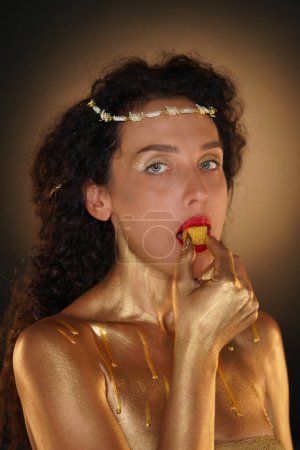 Photo for Beauty and glamour creative advertisement concept. Portrait of female model in studio. Attractive girl wearing tiara and red lipstick, covered in golden paint looking at camera holding candy in lips. - Royalty Free Image