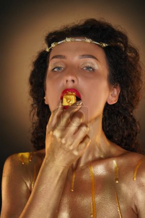 Photo for Beauty and glamour creative advertisement concept. Portrait of female model in studio. Attractive girl wearing tiara and red lipstick, covered in golden paint looking at camera biting golden candy. - Royalty Free Image