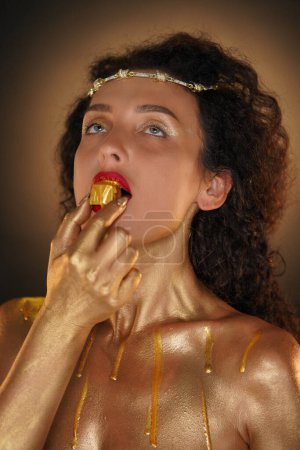 Photo for Beauty and glamour creative advertisement concept. Portrait of female model in studio. Attractive girl wearing tiara and red lipstick, body covered in golden paint holding candy in mouth looking up. - Royalty Free Image