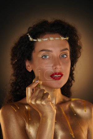 Photo for Beauty and glamour creative advertisement concept. Portrait of female model in studio. Appealing girl wearing tiara and red lipstick, body covered in golden paint holding golden strawberry in hand. - Royalty Free Image