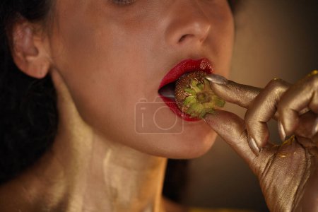 Photo for Beauty and glamour creative advertisement concept. Portrait of female model in studio. Close up of appealing girl face part with red lipstick, neck covered in golden paint holds strawberry in lips. - Royalty Free Image