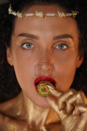 Photo for Beauty and glamour creative advertisement concept. Portrait of female model in studio. Close up face shot girl with red lipstick, covered in golden paint holds strawberry in lips looks at camera. - Royalty Free Image