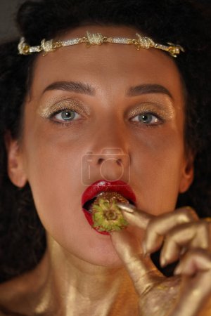 Photo for Beauty and glamour creative advertisement concept. Portrait of female model in studio. Close up face shot woman with red lipstick, coated in golden paint holds strawberry in lips looks at camera. - Royalty Free Image