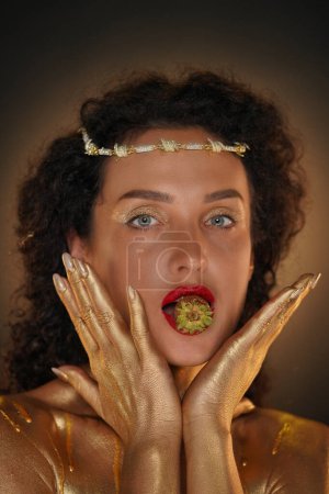 Photo for Beauty and glamour creative advertisement concept. Portrait of female model in studio. Appealing girl in tiara covered in golden paint holds strawberry in lips, hand touching face looks at camera. - Royalty Free Image