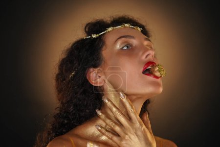 Photo for Beauty and glamour creative advertisement concept. Portrait of female model in studio. Close shot girl in tiara covered in golden paint holds strawberry in lips, hand touching neck, looks away. - Royalty Free Image