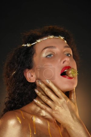 Photo for Beauty and glamour creative advertisement concept. Portrait of female model in studio. Close shot pretty girl in tiara body covered in golden paint with strawberry in lips, hand touching face skin. - Royalty Free Image