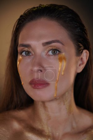 Photo for Beauty and fashion creative advertisement concept. Portrait of brunette female model in studio. Attractive woman with long hair and makeup looking at camera with tears of golden paint flowing down. - Royalty Free Image