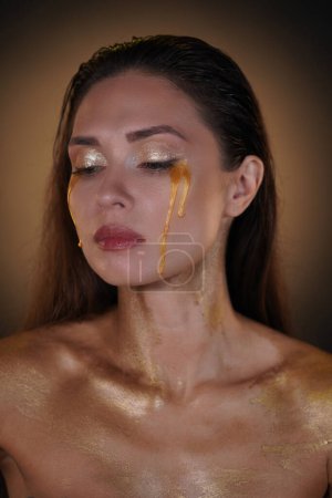 Photo for Beauty and fashion creative advertisement concept. Portrait of brunette female model in studio. Appealing woman with long hair and makeup posing at camera with tears of golden paint flowing down. - Royalty Free Image