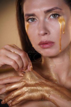 Téléchargez les photos : Concept de publicité créative beauté et mode. Portrait de mannequin femme brune en studio. Belle femme avec des larmes d'or et les mains couvertes de peinture dorée, décoller revêtement d'or sec. - en image libre de droit