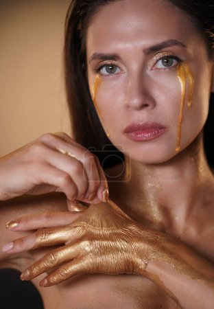 Photo for Beauty and fashion creative advertisement concept. Portrait of brunette female model in studio. Attractive woman with golden tears and fingers covered in golden paint, taking off dry gold coating. - Royalty Free Image