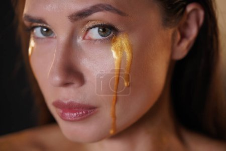Photo for Beauty and fashion creative advertisement concept. Portrait of brunette female model in studio. Attractive woman with makeup looking at the camera with tears of golden paint flowing down the face. - Royalty Free Image
