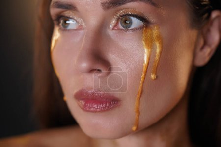 Photo for Beauty and fashion creative advertisement concept. Portrait of brunette female model in studio. Attractive woman with makeup looking sideway from camera with tears of golden paint flowing down. - Royalty Free Image
