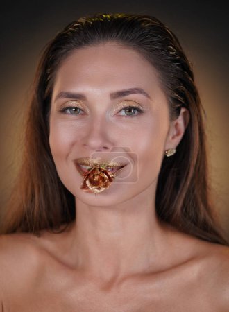 Téléchargez les photos : Concept de publicité créative beauté et mode. Portrait de mannequin femme brune en studio. Femme attrayante avec un maquillage de fard à paupières lumineux tenant fleur de rose dorée dans la bouche souriant à la caméra. - en image libre de droit