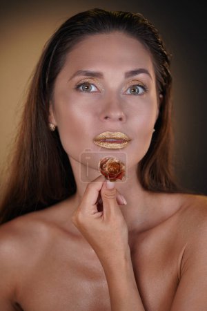Téléchargez les photos : Concept de publicité créative beauté et mode. Portrait de mannequin femme brune en studio. Belle femme avec coiffure et maquillage lumineux tenant fleur de rose dorée regardant la caméra. - en image libre de droit