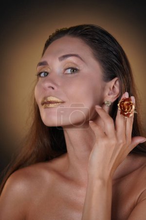 Photo for Beauty and fashion creative advertisement concept. Portrait of brunette female model in studio. Attractive woman with gold lipstick and eyeshadows posing with golden rose flower smiling at the camera. - Royalty Free Image