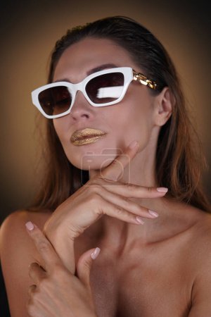 Foto de Concepto de publicidad creativa de belleza y moda. Retrato de modelo femenina morena en estudio. Mujer glamorosa con elegante maquillaje brillante posando en gafas de sol blancas, mirando a la cámara. - Imagen libre de derechos