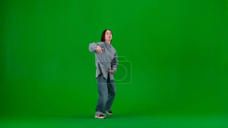 Photo for In the frame on a green background, a limp. Dances young, beautiful girl. Demonstrates dance moves in the style of hip hop. Shes staring at the camera. Shes dressed in loose clothes, street style. - Royalty Free Image