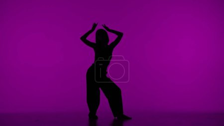 Téléchargez les photos : Dans le cadre sur le fond violet dans la silhouette. Danses élancées, belle fille. Démontre des mouvements de danse dans le style du hip hop, levant les mains vers le haut. C'est féminin, plastique, rythmique. - en image libre de droit