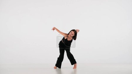 Photo for In the frame on a white background. A young, beautiful girl dances. Demonstrates dance moves in the style of hip hop. She gazes at the camera. Shes feminine, barefoot in a black top and pants. - Royalty Free Image