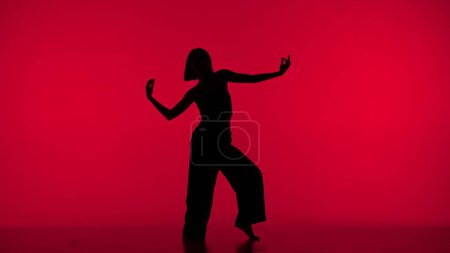 Téléchargez les photos : Dans le cadre sur fond rouge dans la silhouette. Danses élancées, belle fille. Démontre des mouvements de danse dans le style du hip hop. C'est féminin, plastique, rythmique. Plan général. - en image libre de droit