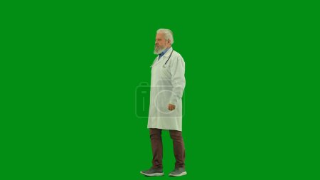 Téléchargez les photos : Santé et bien-être concept créatif. Portrait d'homme médecin en studio sur écran vert chromatique. Médecin principal en uniforme blanc marchant en regardant le visage confiant de la caméra. Vue du profil. - en image libre de droit