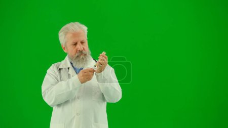 Photo for Healthcare and wellness concept. Portrait of man medic in studio on chroma key green screen. Senior doctor close up in white uniform standing draws injection in syringe, holding in hand. Middle shot. - Royalty Free Image